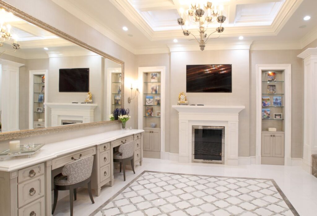 A luxury vanity and dressing room featuring a large, ornate silver-framed mirror over a light-grey double vanity with marble tops and two textured grey chairs.