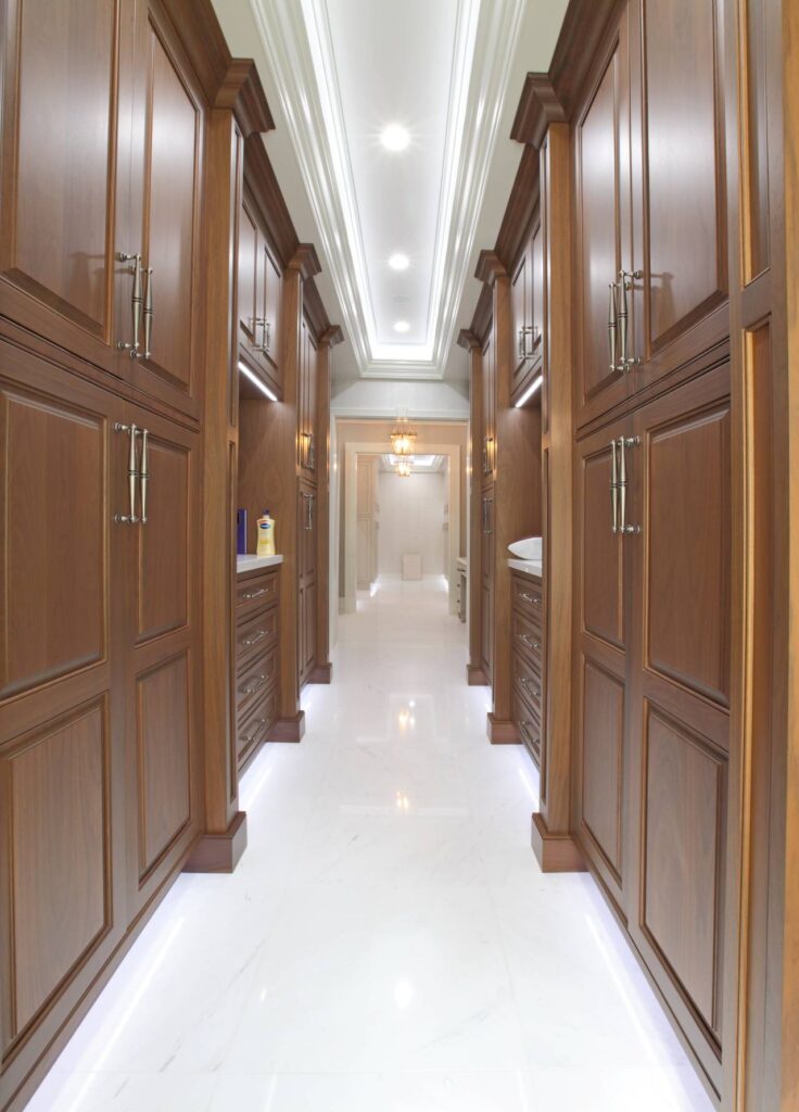 A symmetrical, eye-level view of a luxurious walk-in dressing area or hallway featuring floor-to-ceiling walnut cabinetry with ornate paneling and antique brass hardware. The space is anchored by a polished white marble floor with integrated LED toe-kick lighting that casts a bright glow along the base.