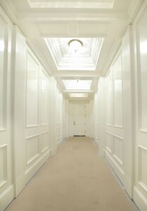A bright, long hallway with ornate white-painted wall paneling and a series of deeply recessed, tiered coffered ceilings. Each ceiling section contains a glowing light fixture, and the floor is covered in a neutral tan protective runner, leading to a closed white door at the far end.