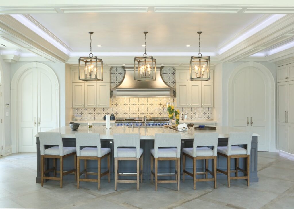A wide-angle, symmetrical shot of a luxury kitchen featuring a large dark-grey island topped with white marble. Six wood-framed counter stools with light-grey upholstery are lined up along the island. Above the island hang three bronze lantern-style chandeliers.