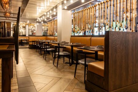 A warm, atmospheric shot of the "All Set" dining room highlighting the chevron-patterned wood floors. The scene features dark wood booths with tan leather upholstery and black dining chairs with blue-and-white patterned seats. In the background, a curtain of vertically hanging ropes with decorative knots serves as a room divider, complemented by a string of Edison bulb pendant lights that cast a soft glow across the industrial-style ceiling.