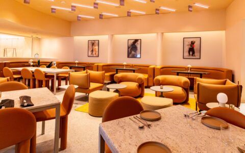 A comprehensive wide-angle shot of the "Manifest" dining room, showcasing a variety of ochre-colored seating options, from curved wall booths to plush, low-slung armchairs and cylindrical ottomans. The room features light granite-topped tables and a unique ceiling installation of suspended, linear LED lights. The warm, monochromatic color palette is reinforced by recessed cove lighting and a series of framed abstract artworks along the far wall.